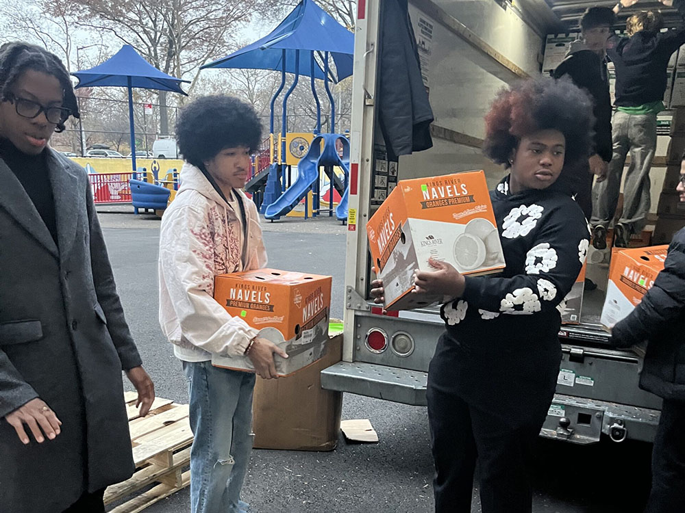 Volunteers unload orange Navels juice boxes from a delivery truck in a parking lot near a playground helping move supplies