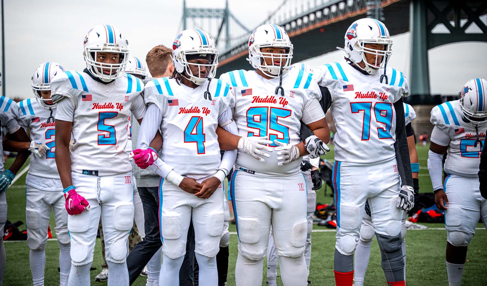 Football team in white uniforms with blue accents huddles on a field ready for play under a bridge in the background