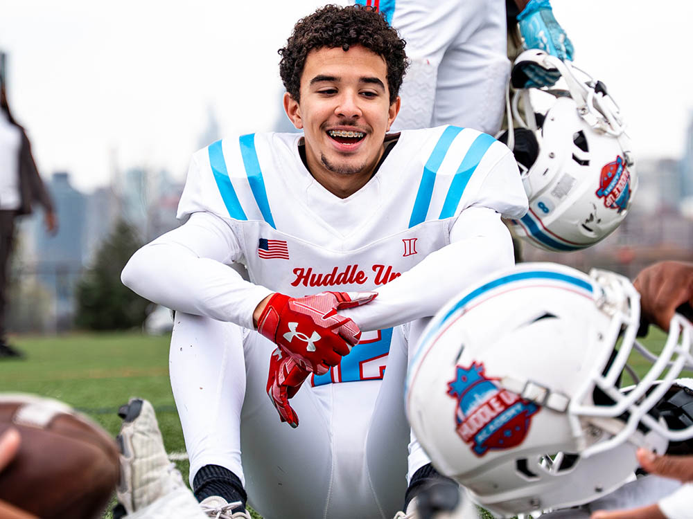 Smiling football player in a white and blue uniform sits on the grass with red gloves helmet nearby